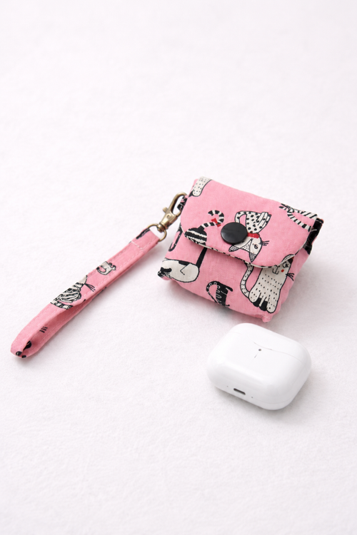 A pink fabric AirPods case with a black and white animal print pattern, featuring a matching wrist strap and a snap button closure, shown next to a white AirPods charging case on a white background.