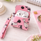 Pink fabric AirPods case with a cat print pattern, shown open with white AirPods inside on a light tabletop. The case has black buttons, a wrist strap, and a metal clasp, with nearby stationery props including a pink notebook and flowers.