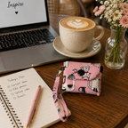 A pink patterned crossbody phone pouch with a wrist strap sits on a wooden table next to an open laptop, a cup of latte, and a notebook in a cafe setting.
