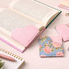 Three fabric bookmark holders with heart-shaped cutouts are displayed on a light wood desk. Two are pink with white polka dots, and one is a blue floral print with pink roses.