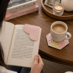 A person holds an open book on a wooden table, with a pink polka-dot bookmark tucked into the page. A cup of coffee, a candle, and stacked books are arranged around it in a cozy indoor setting.