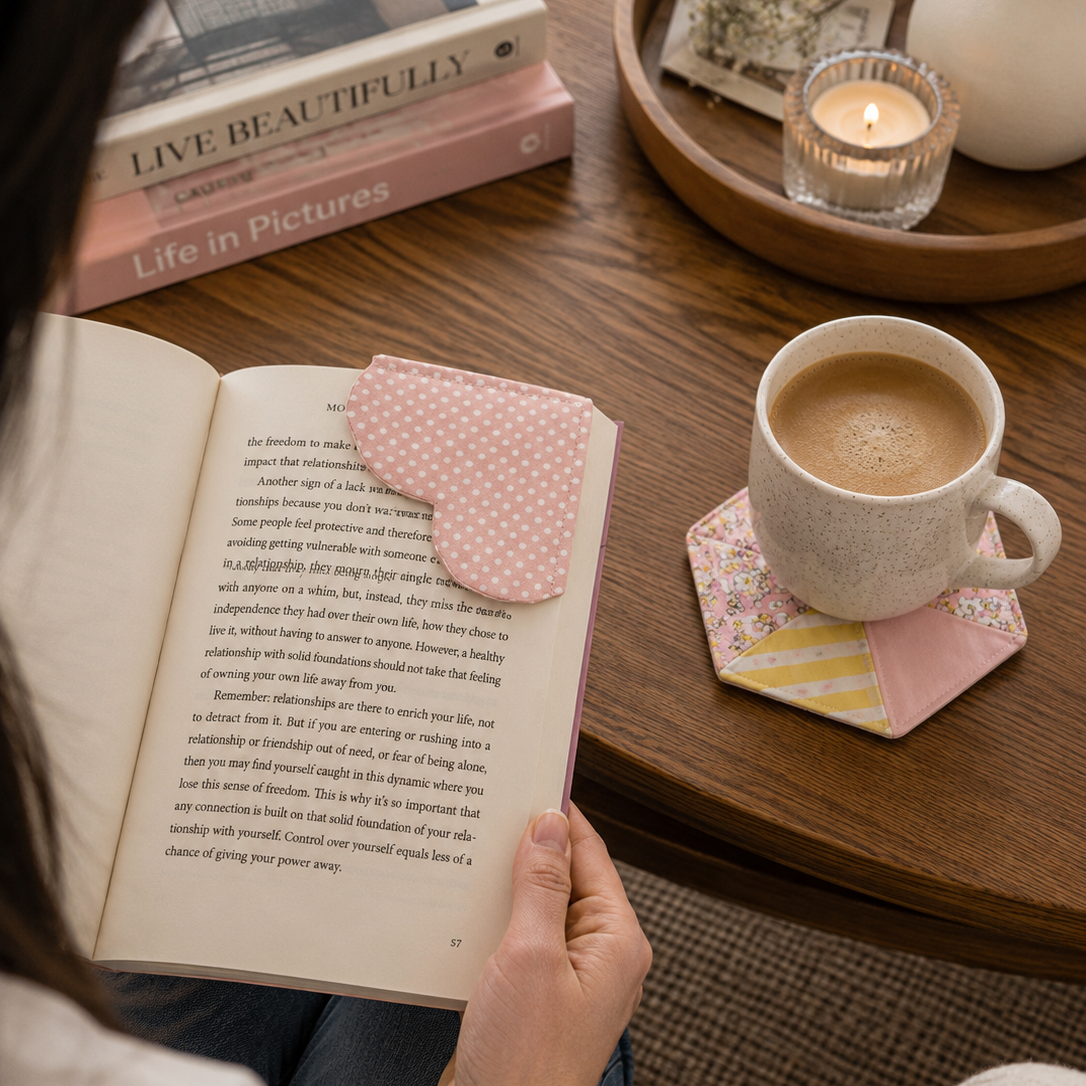 A person holds an open book on a wooden table, with a pink polka-dot bookmark tucked into the page. A cup of coffee, a candle, and stacked books are arranged around it in a cozy indoor setting.