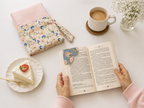 A top-down lifestyle scene showing a person's hands holding an open book with a floral bookmark. To the left is a pink striped and floral fabric book pouch, a slice of cake on a plate, a cup of coffee, and a vase of white flowers on a white table.