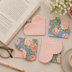 Four fabric book coasters arranged on a light tabletop with styled props, including an open book, eyeglasses, a pink pen, a cup of coffee, and flowers. Two coasters are pink with white polka dots and two are blue with a pink rose floral print.