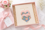 A gift set featuring a book titled 'The Little Book of HAPPINESS' with three heart-shaped fabric covers in pink, blue, and floral print, presented in a pink box with shredded paper. A second wrapped gift box with a pink ribbon and card reading 'Especially For You' sits beside it on a light marble surface with flowers and a card in the background.