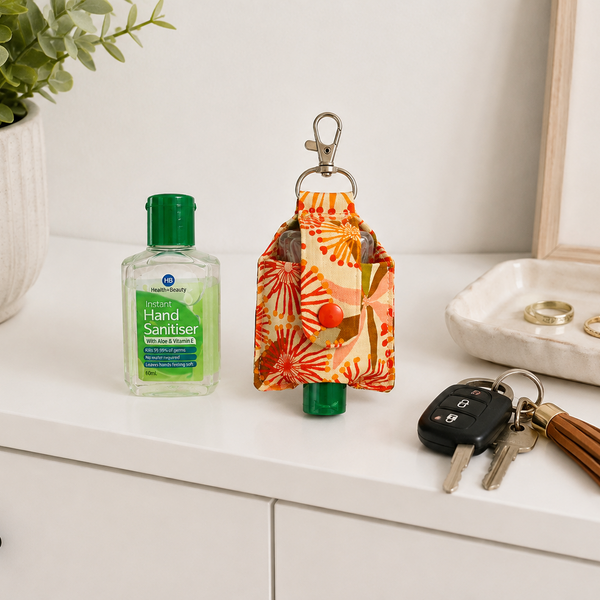A small fabric key pouch with a green cap and orange floral print, shown on a white dresser alongside a hand sanitizer bottle, keys, and a decorative tray in a bright indoor setting.