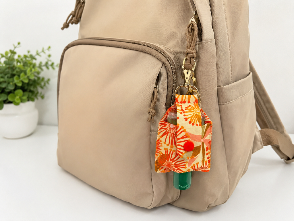 A beige nylon backpack with a side pocket, shown with a small orange and yellow floral patterned hand sanitizer pouch hanging from a gold zipper pull.