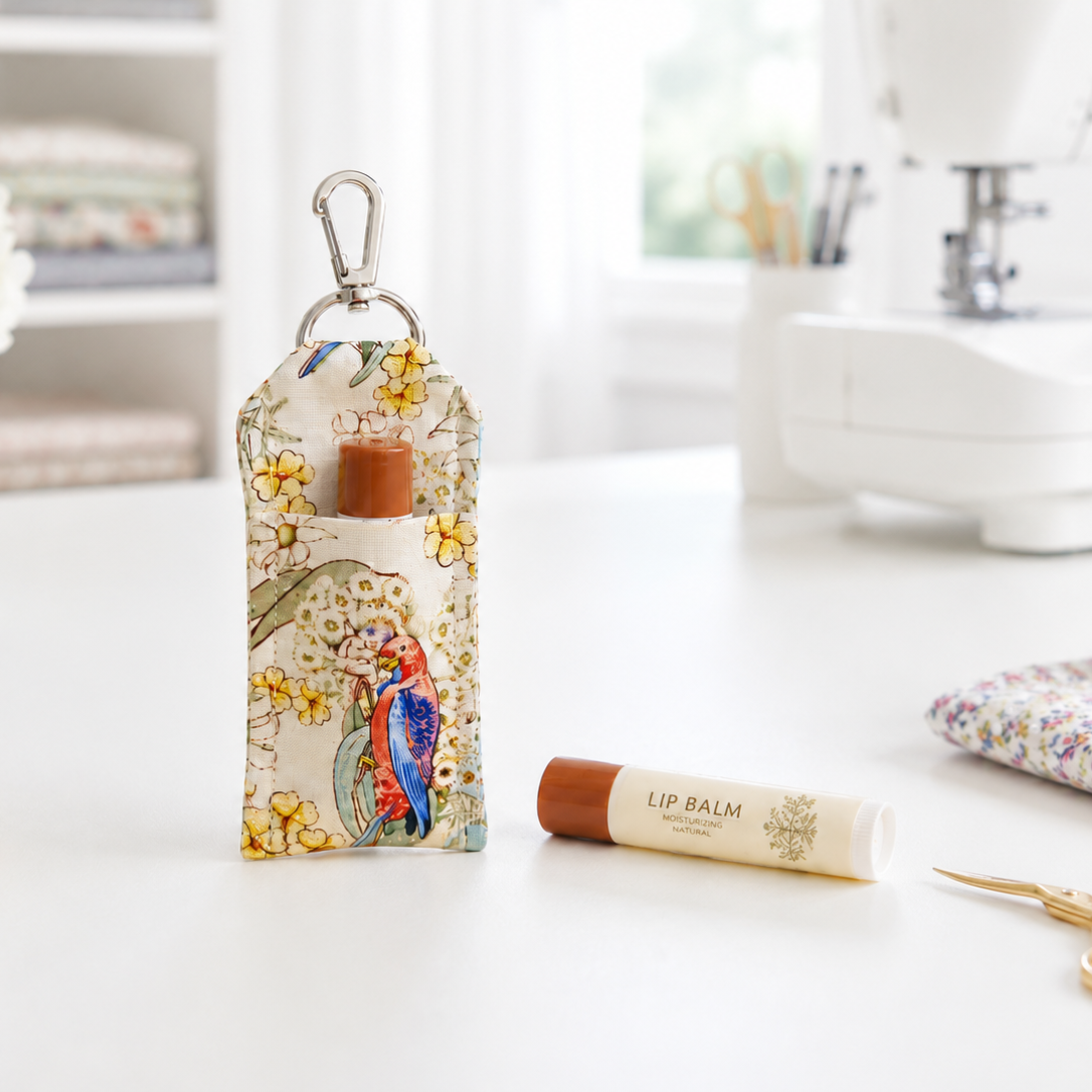 A small fabric lip balm pouch with a colorful bird and floral print stands on a white desk, holding a brown lip balm tube. A matching lip balm tube lies beside it, with sewing-themed props like scissors, a pincushion, thread, and a sewing machine blurred in the background.