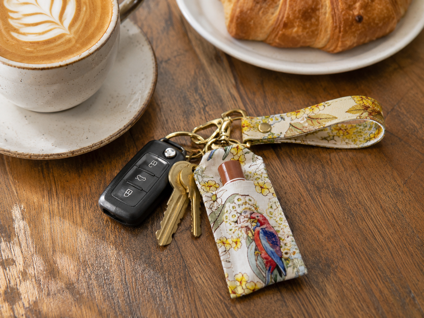 A fabric keychain pouch with a floral print and a colorful bird illustration, attached to gold keys and a black car key fob, resting on a wooden table next to a cup of coffee and a croissant.