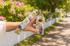 A hand holding a matching fabric wristlet strap and small card holder with a colorful bird and floral print, photographed outdoors on a sunny path with flowers and greenery in the background.