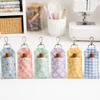 Six fabric lip balm holders with metal clips are lined up on a white tabletop. Each pouch has a different pastel floral or gingham pattern: blue daisies, pink daisies, mint daisies, lavender daisies, yellow daisies, and blue-and-white gingham.
