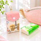 A pink and white striped fabric pouch with a zipper lies on a white tabletop next to a small printed fabric card holder with a Eiffel Tower design and a green tissue pack. A potted plant and a framed heart print are softly blurred in the background.