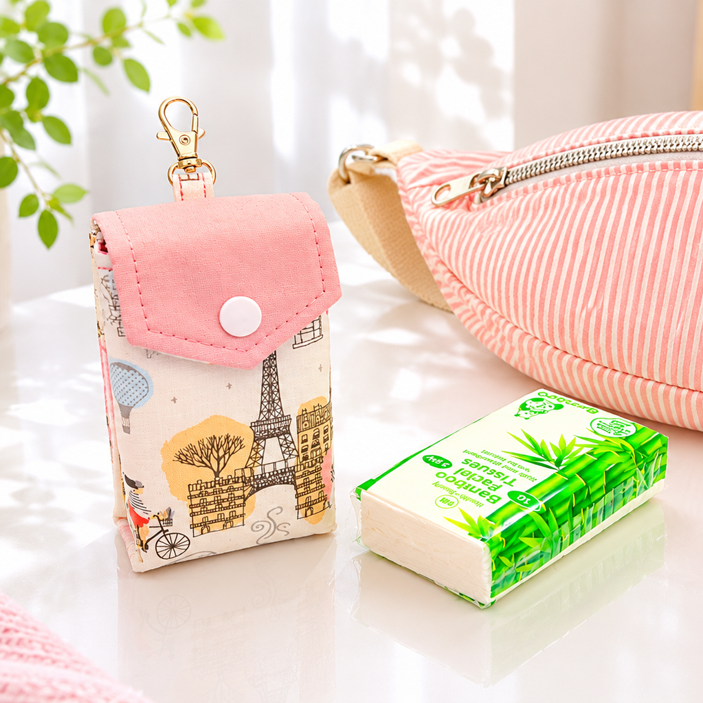 A pink and white striped fabric pouch with a zipper lies on a white tabletop next to a small printed fabric card holder with a Eiffel Tower design and a green tissue pack. A potted plant and a framed heart print are softly blurred in the background.