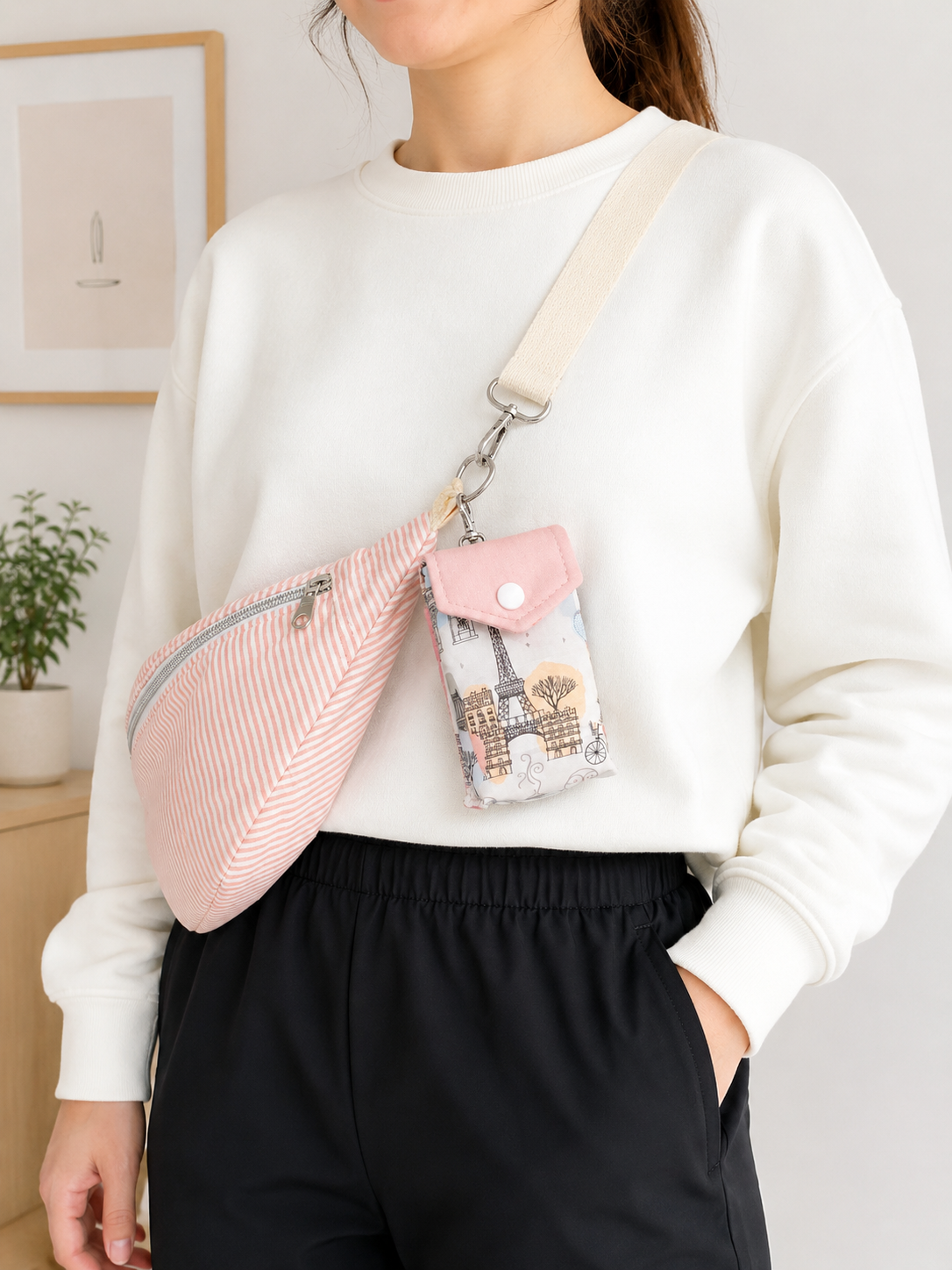A woman wearing a white sweatshirt and black pants carries a pink and white striped sling bag with a matching small pouch featuring a Paris Eiffel Tower print.