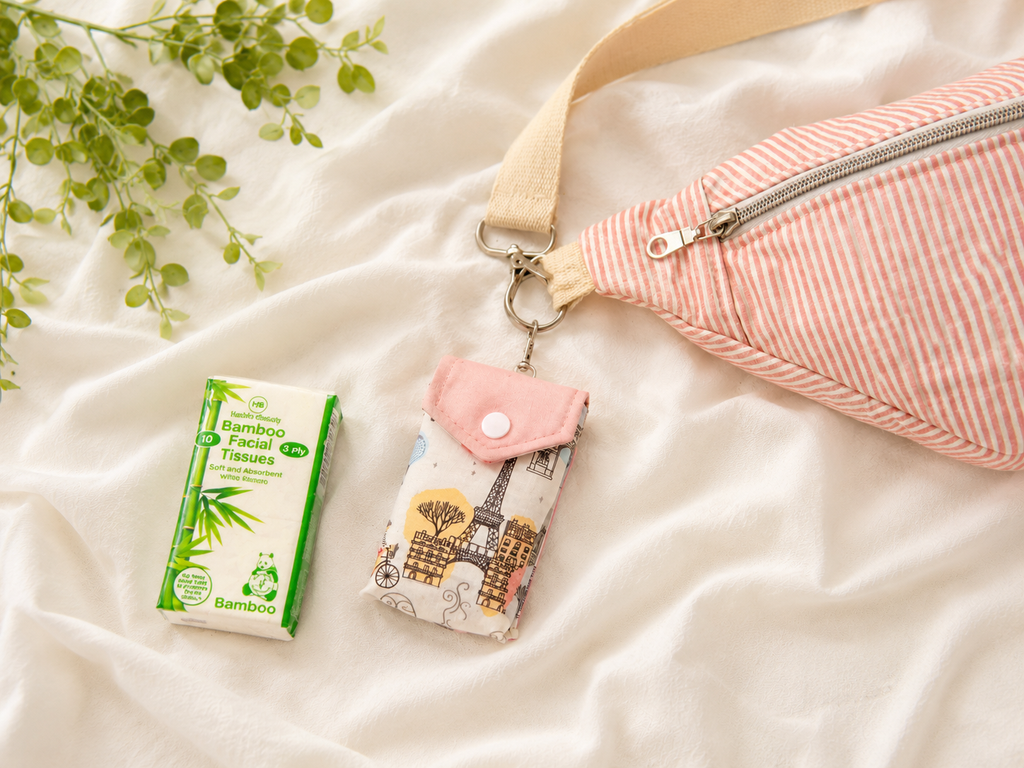 A pink and white striped fanny pack with a cream strap is styled on white fabric alongside a Paris-themed facial tissue pouch and a pack of bamboo facial tissues.